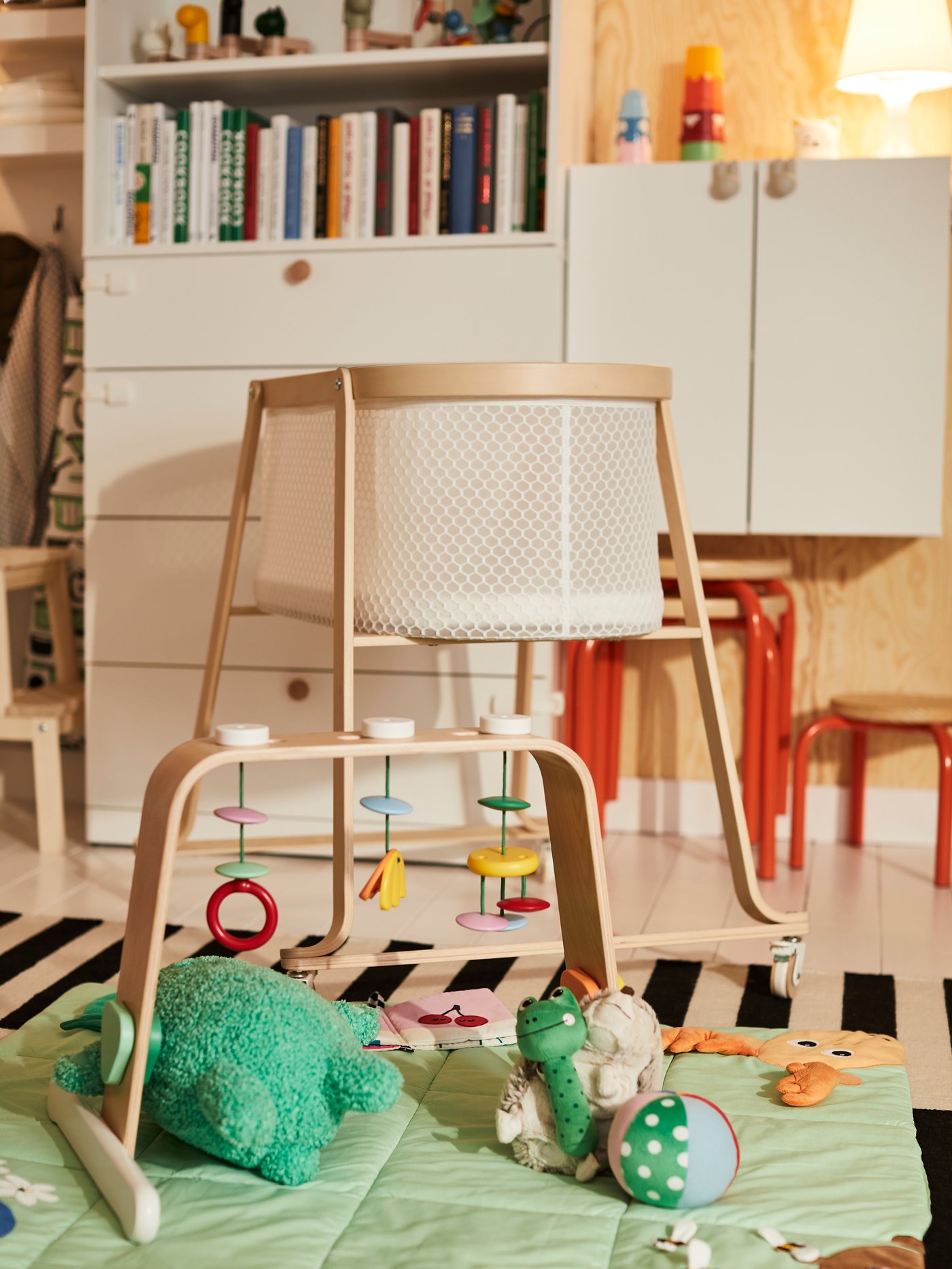 En vit ÄLSKVÄRD babysäng med träram står i ett barnrum, omgiven av leksaker på en lekmatta och möbler i ljusa färger.