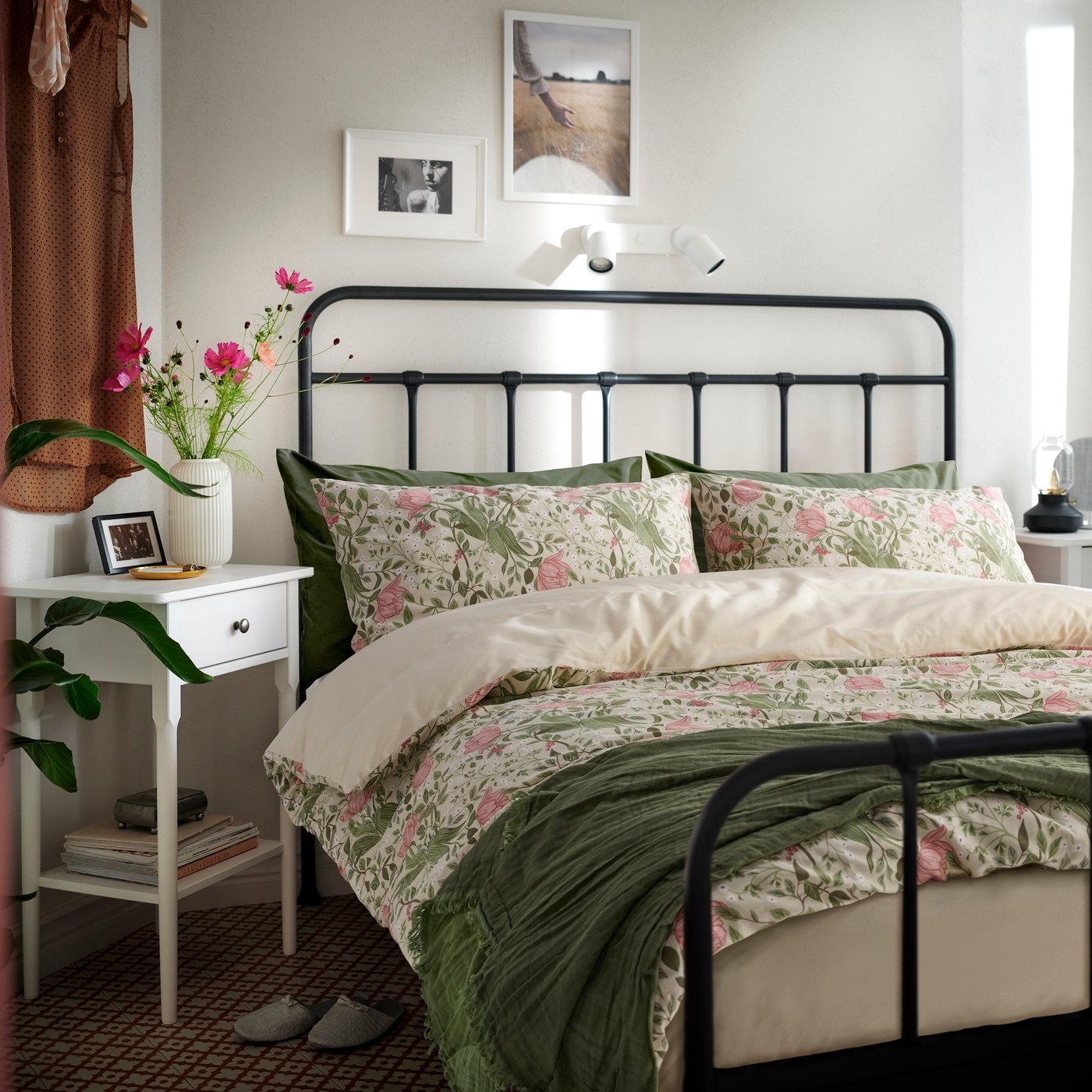 Уютная спальня с черной металлической кроватью и постельным бельем с цветочным узором в нежных зеленых и розовых тонах, дополненным кремовым пуховым одеялом и зеленым пледом. На белой прикроватной тумбочке стоит ваза со свежими цветами, небольшая фотография в рамке и лампа. Нейтральные стены украшены простыми картинами в рамах, а окно с теплыми шторами пропускает естественный свет, создавая спокойную и уютную атмосферу.

