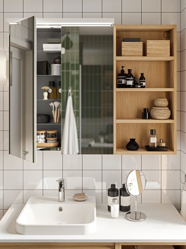 LETTAN medicine cabinet open to show shelves with small bottles. Open shelving on the right with small storage boxes and bottles. Round cosmetic mirror on countertop