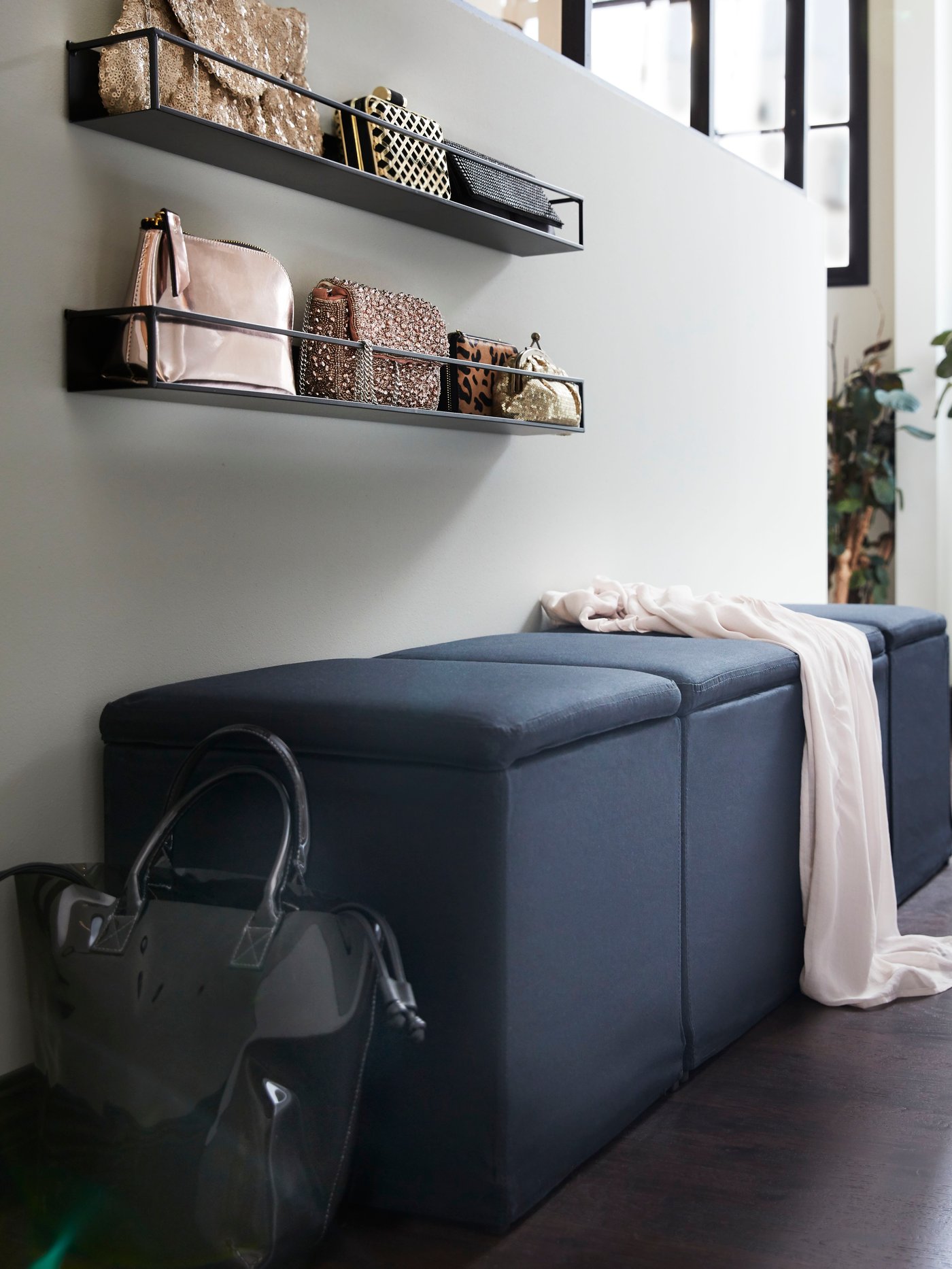 Czarny podnóżek ze schowkiem, czarne półki z torbami i zielona roślina.