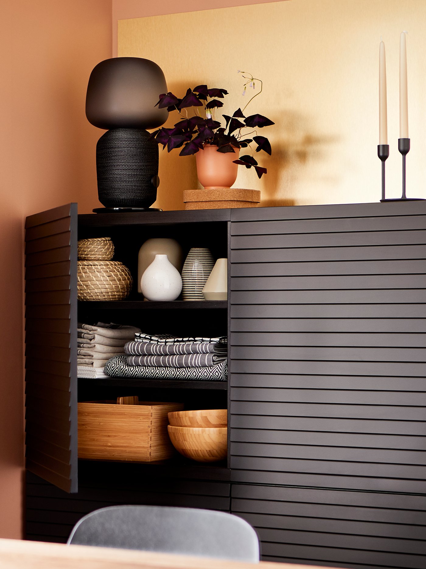 Mueble modular BESTÅ negro con una puerta abierta que muestra mantelería, jarrones y pocillos, con una lámpara/parlante inteligente en la parte superior.