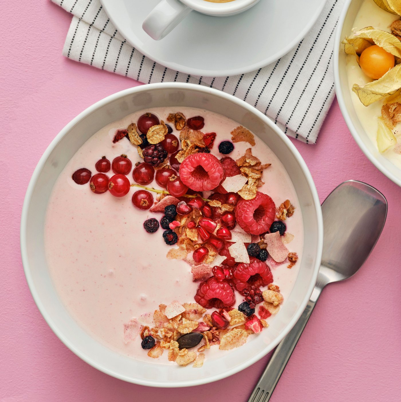 A white bowl has a mix of yoghurt and LÅNGLUR fruit smoothie and it’s decorated with red berries and HJÄLTEROLL granola.
