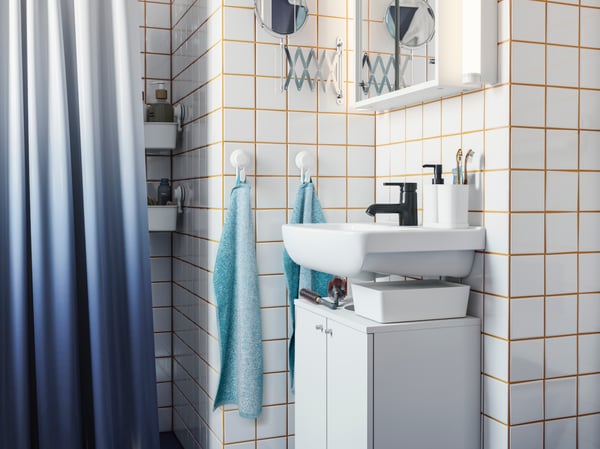Baño pequeño con los muros cubiertos de cerámicas cuadradas blancas y fragüe amarillo, destacan un pequeño vanitorio separado de un mueble blanco que va abajo, dos toallas de mano celestes y una cortina de ducha en degradé blanco a azul. 
