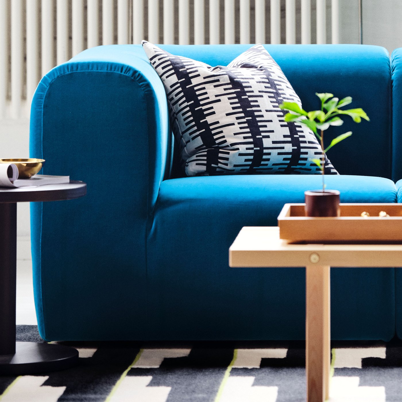En blå STOCKHOLM soffa med ett svartvitt mönstrat kuddfodral står bredvid ett litet sidobord och en träbricka med en liten planta, på en grafisk mönstrad matta.