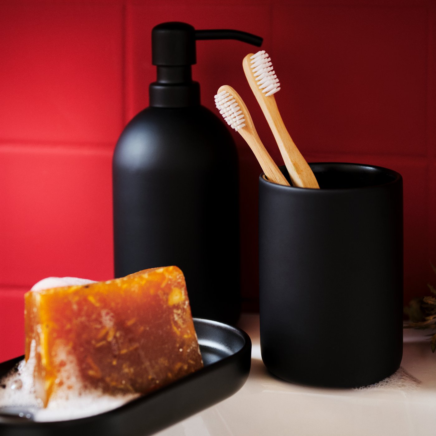 A dark-matte, stoneware GANSJÖN three-piece bathroom set on the side of a white washbasin, next to a red-tiled wall.