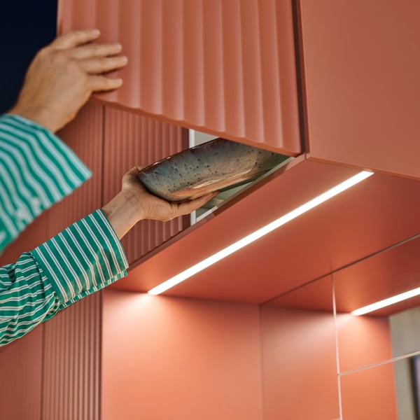 Une personne vêtue d'une chemise à rayures vertes sort un bol d'un placard dont la porte est décorée d'un motif TERRSJÖ à vagues rouge-brun.