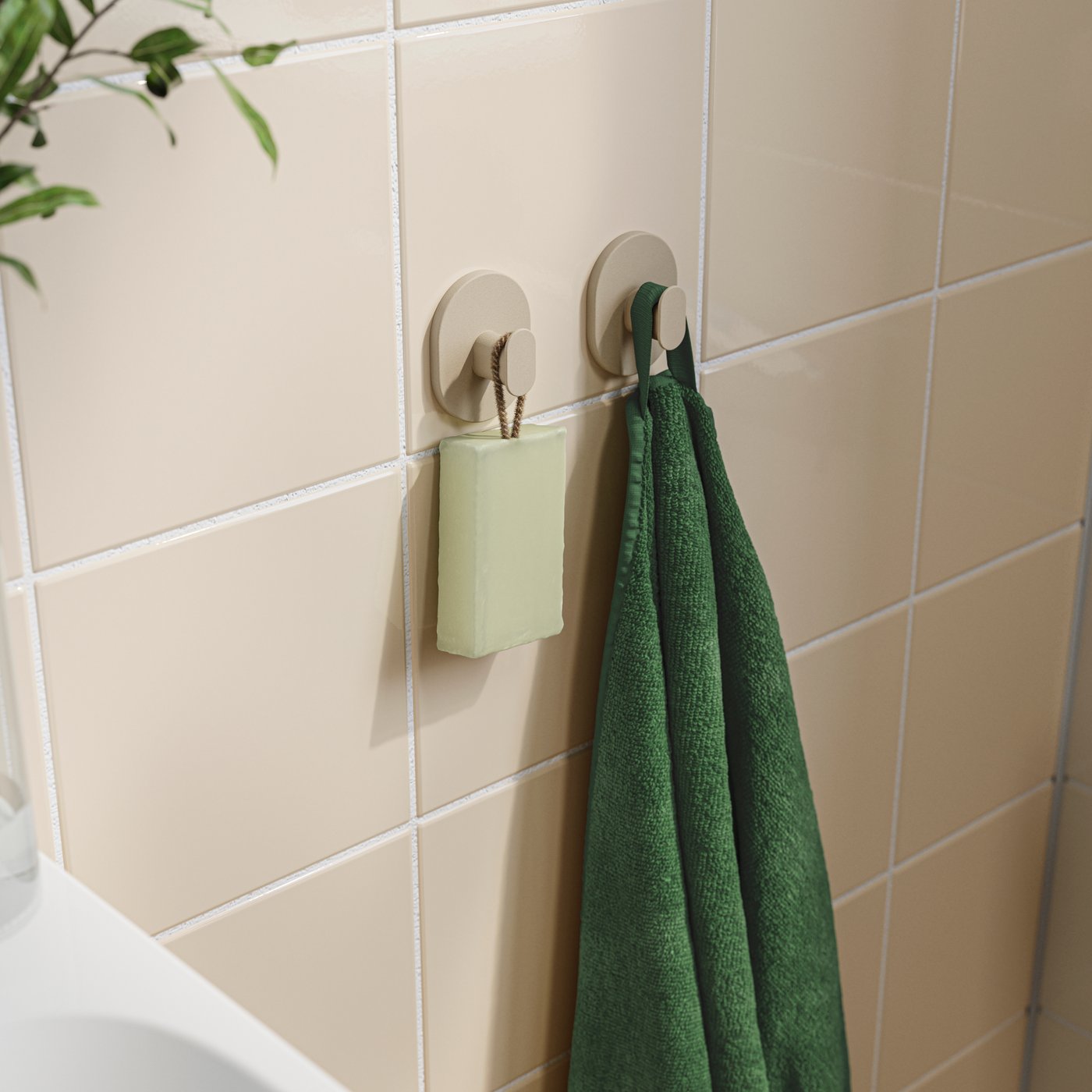 A set of two grey-beige FLORSJÖN hooks in coated metal are on a tiled bathroom wall, holding a green towel and a bar of soap.