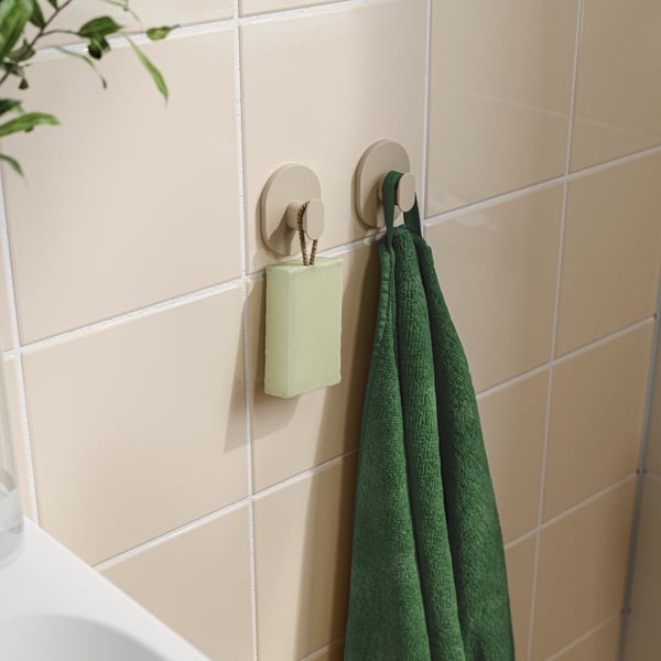 Un ensemble de deux crochets FLORSJÖN gris-beige en métal revêtu est posé sur un mur carrelé de la salle de bain, supportant une serviette verte et un pain de savon.