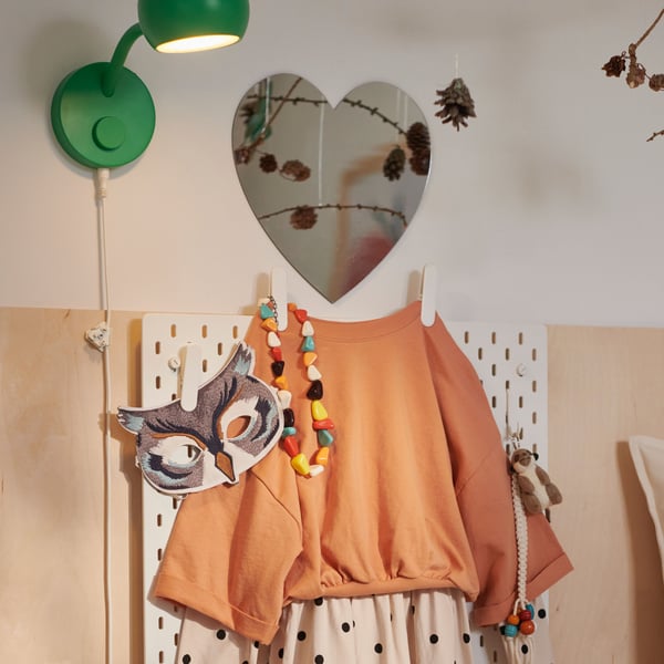 A beige and orange child’s outfit hangs on a SKÅDIS pegboard topped by a HOLMÖARNA heart-shaped mirror.