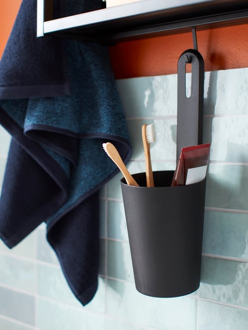 Nahaufnahme eines Bereichs im Badezimmer mit einem SKATTÅN Behälter, in dem Zahnbürsten und Zahnpasta zu sehen sind, an einem ENHET Haken, Fliesen und einem Wandschrank mit Spiegel.