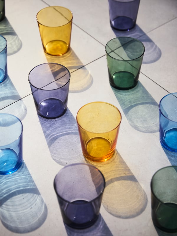 PAPPERSBJÖRK glasses in colourful blue, yellow, green, and purple are displayed on a table, bathed in summer light.