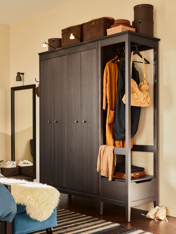 A wooden wardrobe with closed doors and rails on the side with dresses and a bag hanging. Woven boxes sit on top.