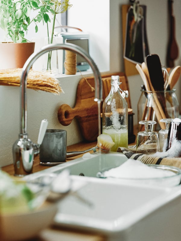Liquid in a clear glass KORKEN bottle with stopper and an oak ARTISTISK chopping board next to a sink of running water.