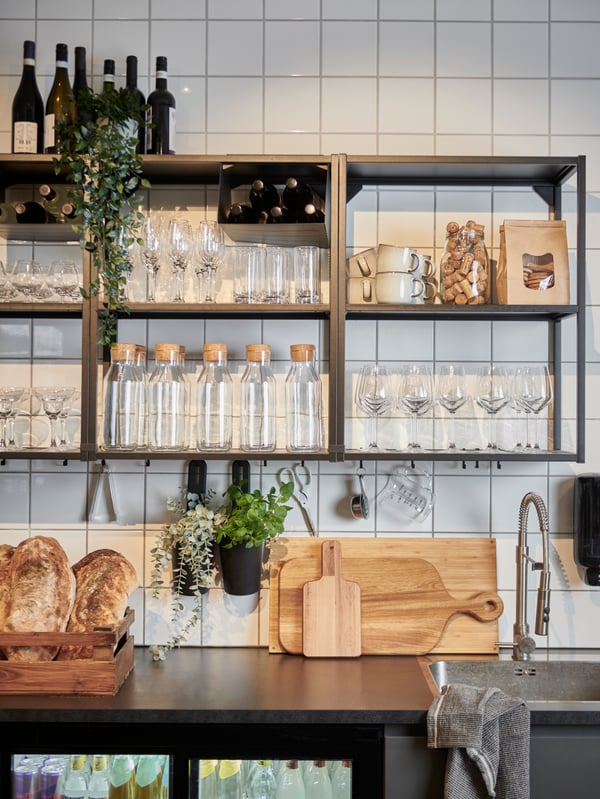 Svart serveringsbänk i ett cafè med diskho och kran. Ovanför, på en vitkaklad vägg, finns flera svarta ENHET hyllor fyllda med glas och muggar.