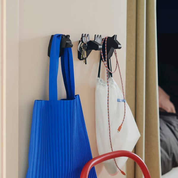 Tre svarta FRÖSÅDD krokar, formade som djurhuvuden, sitter monterade på en vägg med väskor, nycklar och kablar hängande på dem.
