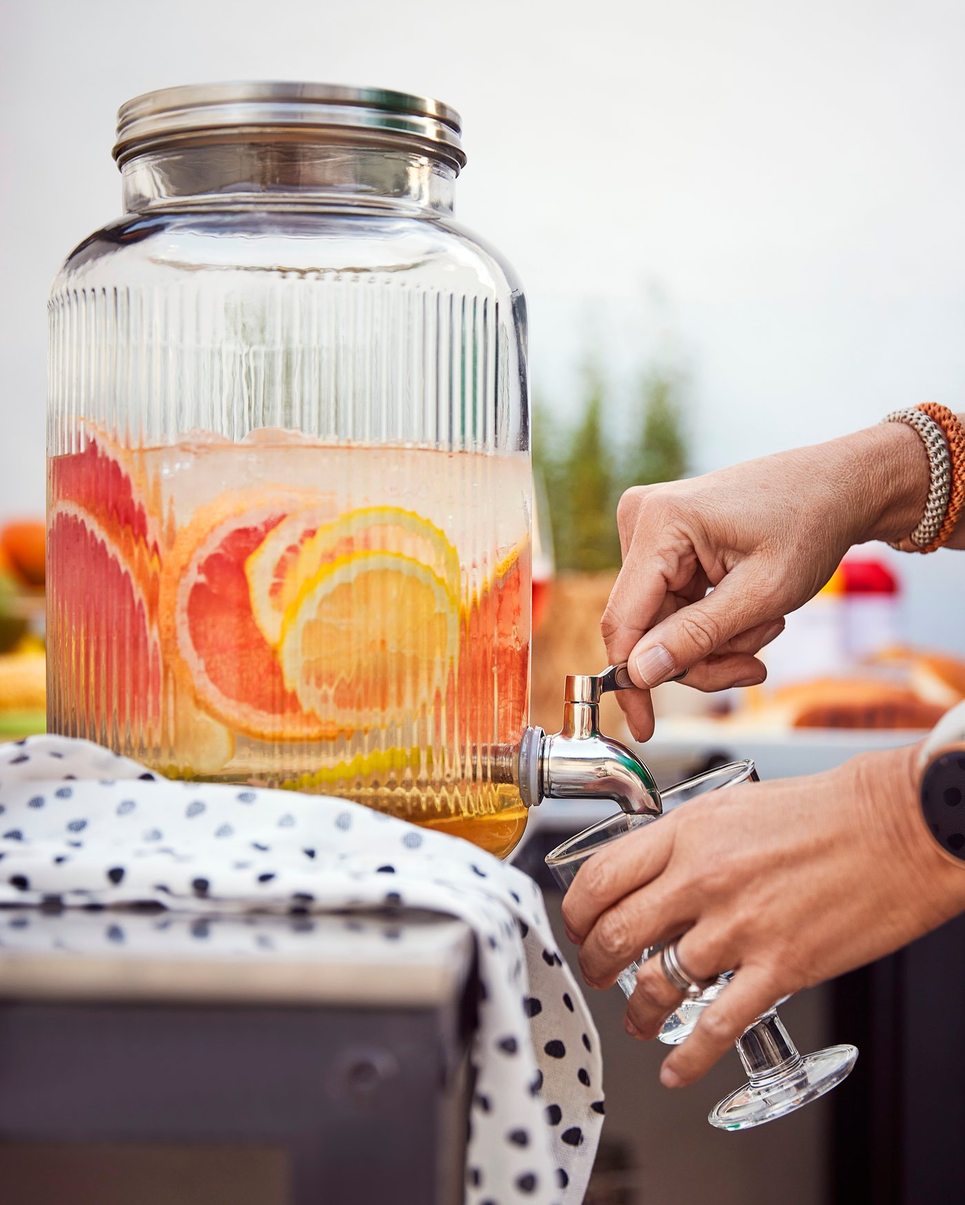 Dispenser de băuturi IKEA din sticlă, plin cu apă infuzată cu felii de citrice colorate, în timp ce o persoană toarnă limonadă rece într-un pahar – perfect pentru petreceri în aer liber.