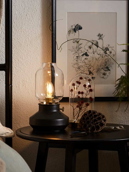 A dark grey metal TÄRNABY table lamp on a bedside table with dried flowers and a framed floral print on the wall behind it.