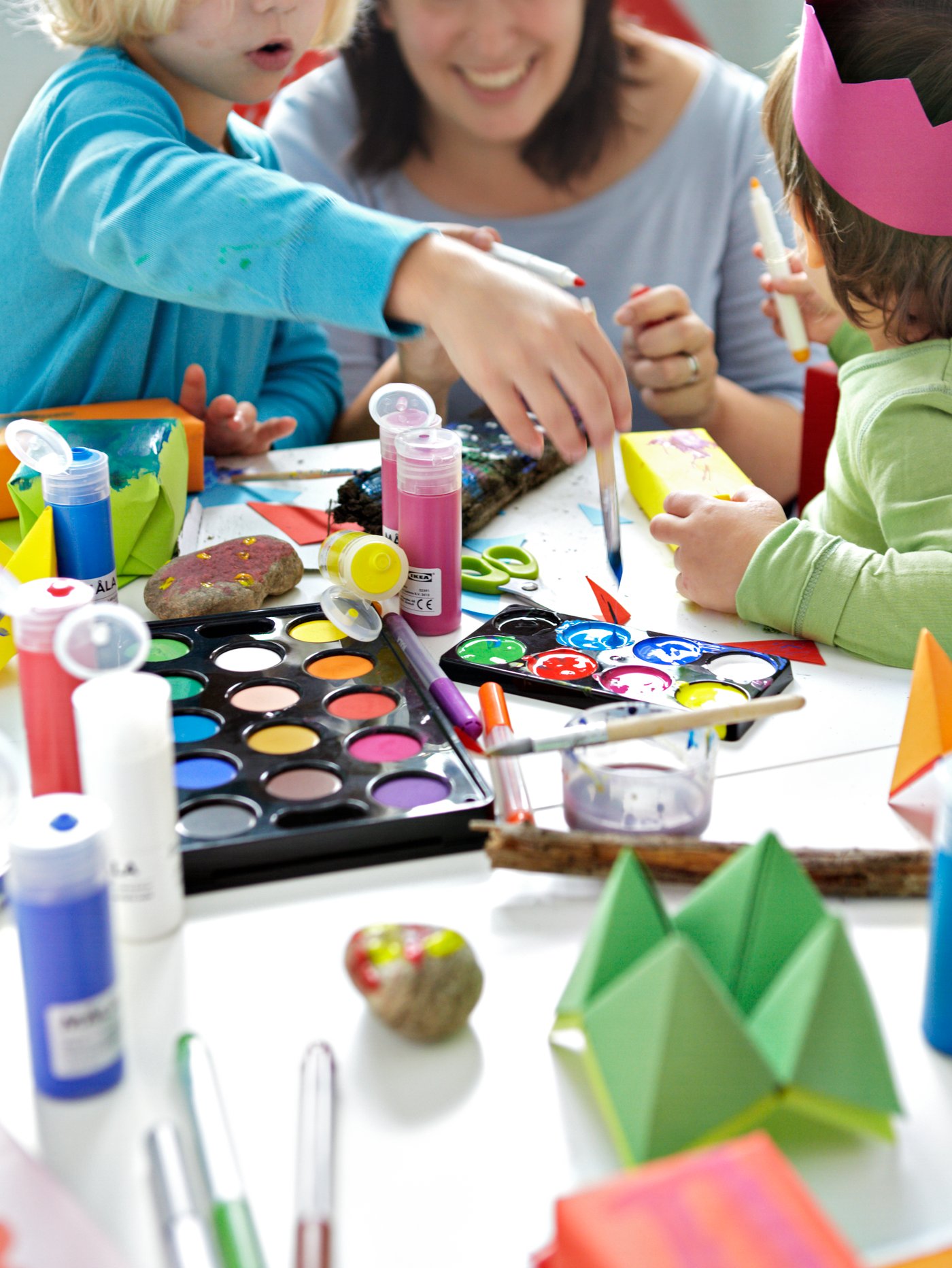 Una mujer y dos niños pintando y dibujando en una mesa cubierta con papel y una bandeja de acuarelas MÅLA, rotuladores y pinturas.