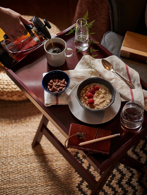 Ein dunkelroter MARYD Tabletttisch auf dem eine Schüssel Porridge mit Himbeeren, eine Schale Nüsse und eine Tasse Kaffee steht.