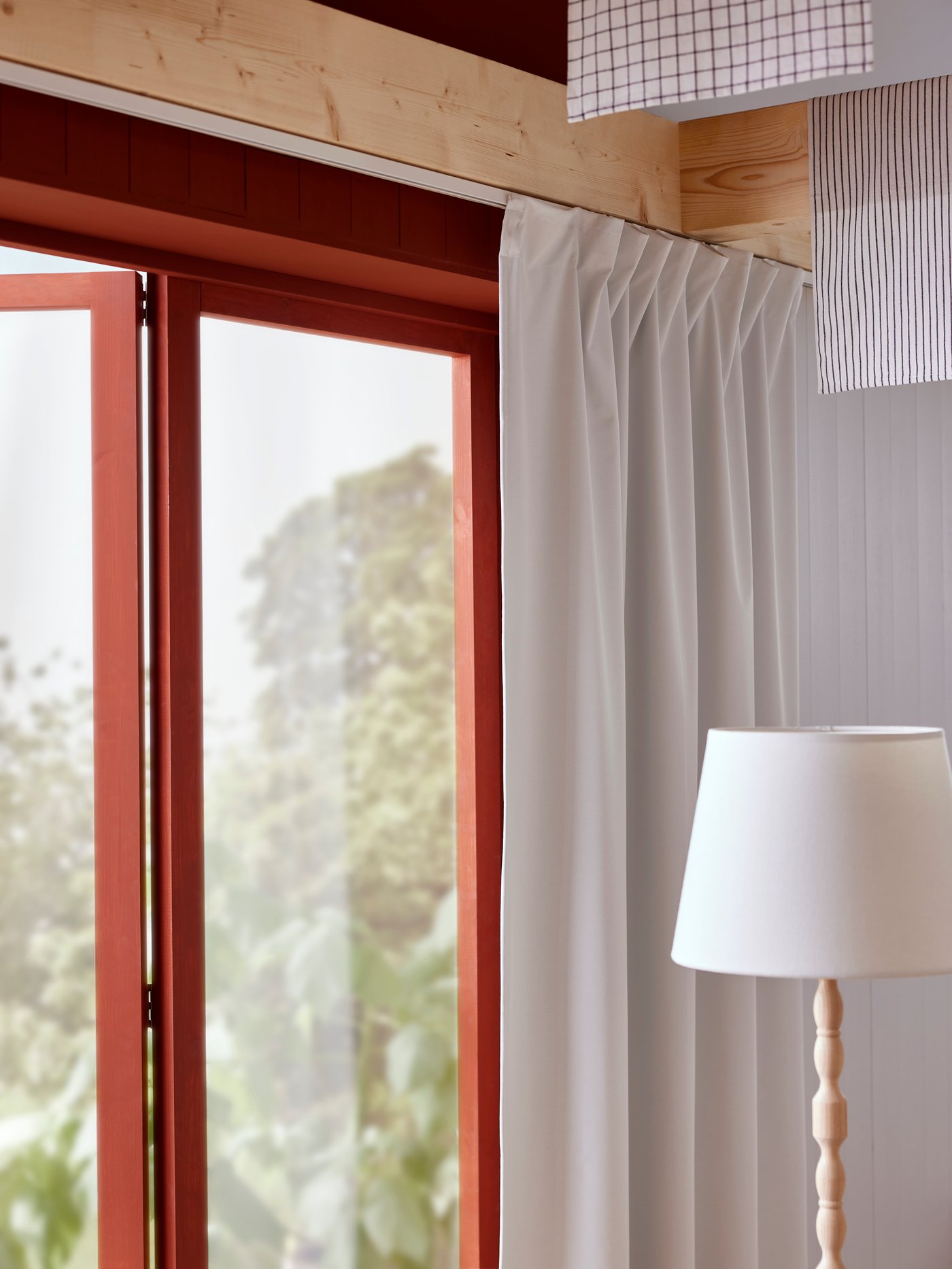 Biała zasłona zaciemniająca KORGMOTT zawieszona na pojedynczej szynie VIDGA przed oknem, przy którym stoi lampa podłogowa KINNAHULT.