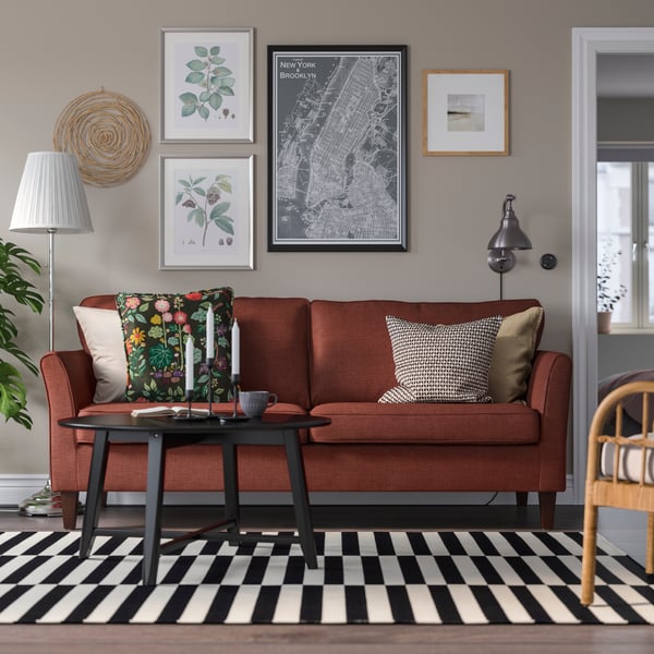 Ciemnoczerwona sofa EKHOLMA stojącej w jasnym pokoju dziennym na tle rbeżowej ściany z galerią obrazków w ramach.