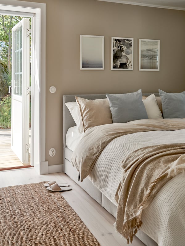 Sovrum med stor säng bäddad i beige och ljusgrått, flera kuddar i matchande toner, tre tavlor ovanför sängen, öppen dörr mot grönskande utomhusmiljö, vävd matta och ett par tofflor på golvet.