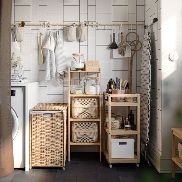 A laundry nook with RÅGRUND trolley, shelves and laundry basket, all in natural materials. Cleaning tools and ironing board.