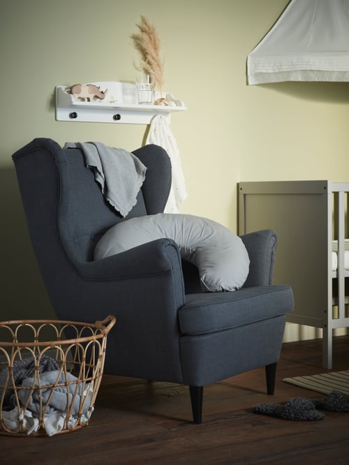 Une chambre d’enfant avec une bergère sur laquelle sont posés un coussin d’allaitement et une couverture, une étagère murale et un lit d’enfant avec un ciel de lit.