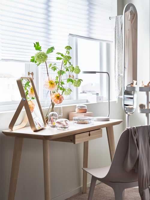 A LISABO desk in ash veneer, with flowers in a vase, mirror and lamp, in front of a window with white blinds.