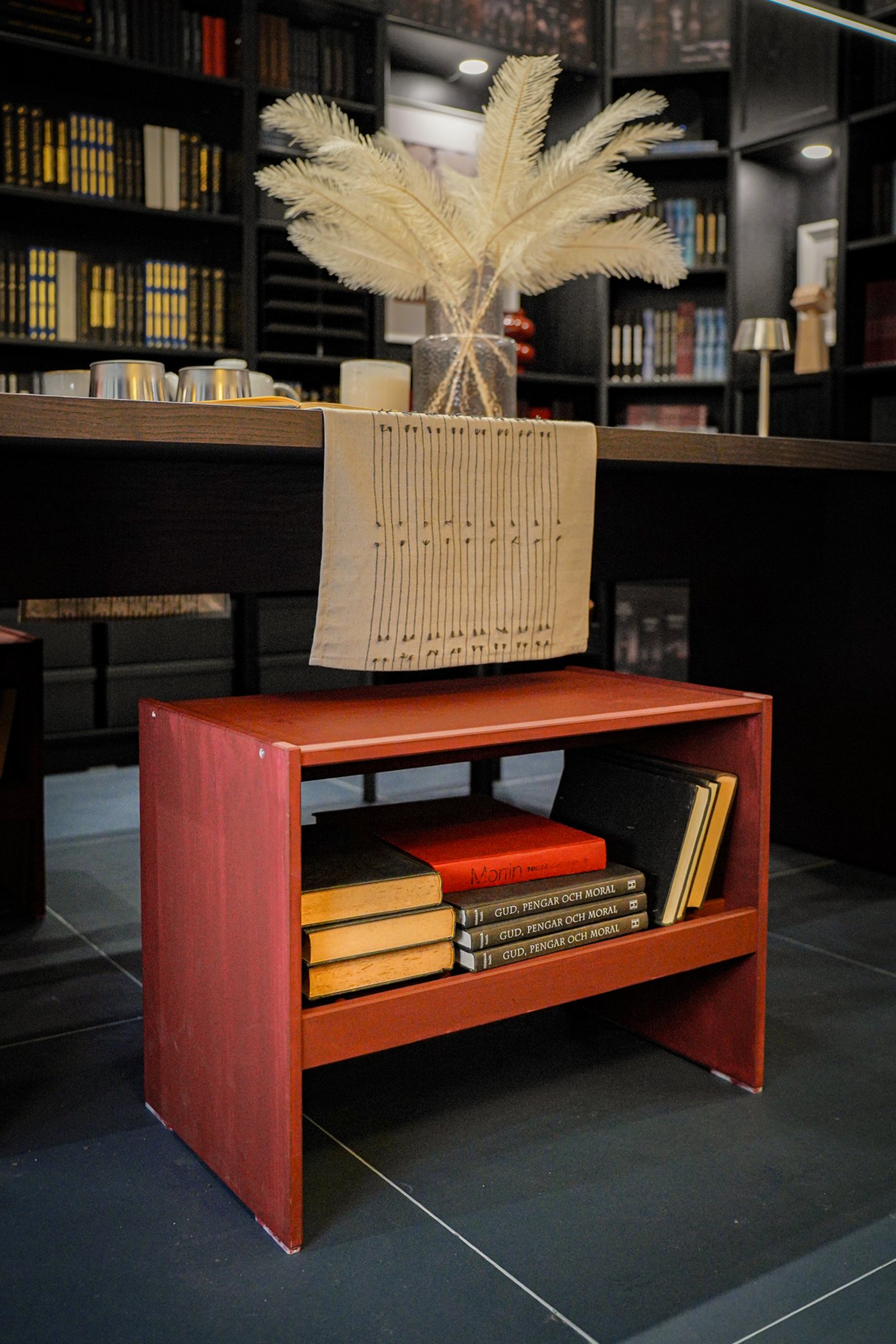 Curious Library room setting at IKEA Tampines, featuring a pine RAGGBERG chest of drawers, painted red, tucked under a dark brown BERGSHYTTAN dining table, against black/brown BILLY bookcases filled with books.