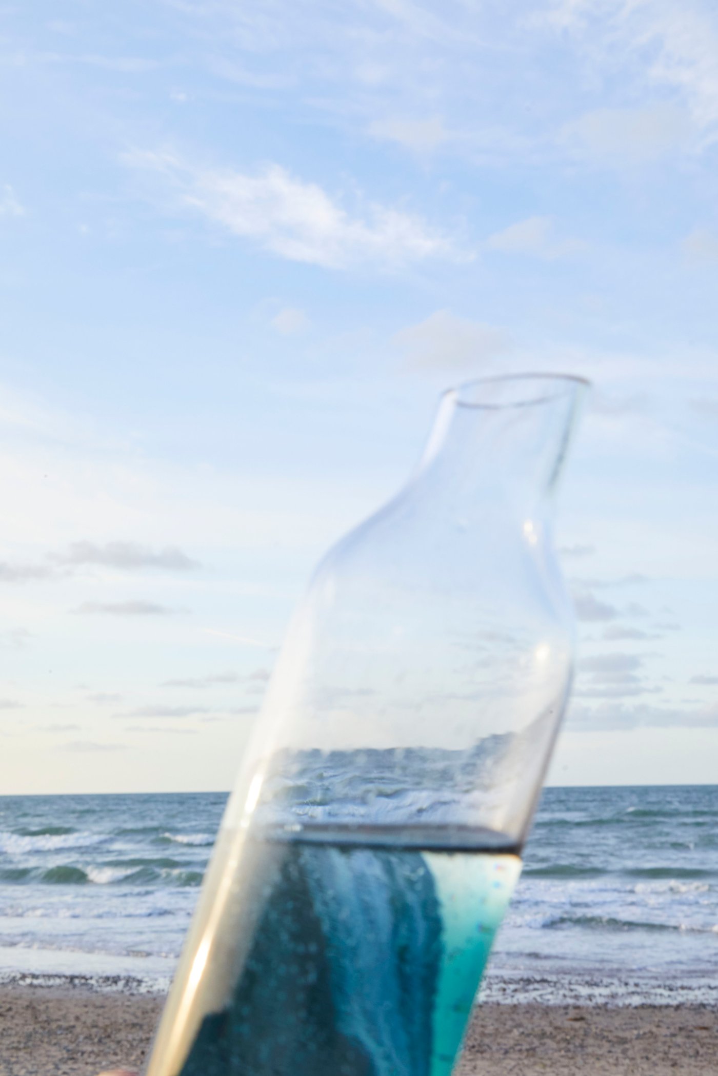Una bottiglia che riflette il mare sui toni dell'azzurro e del blu. Sullo sfondo, un cielo azzurro e il mare.