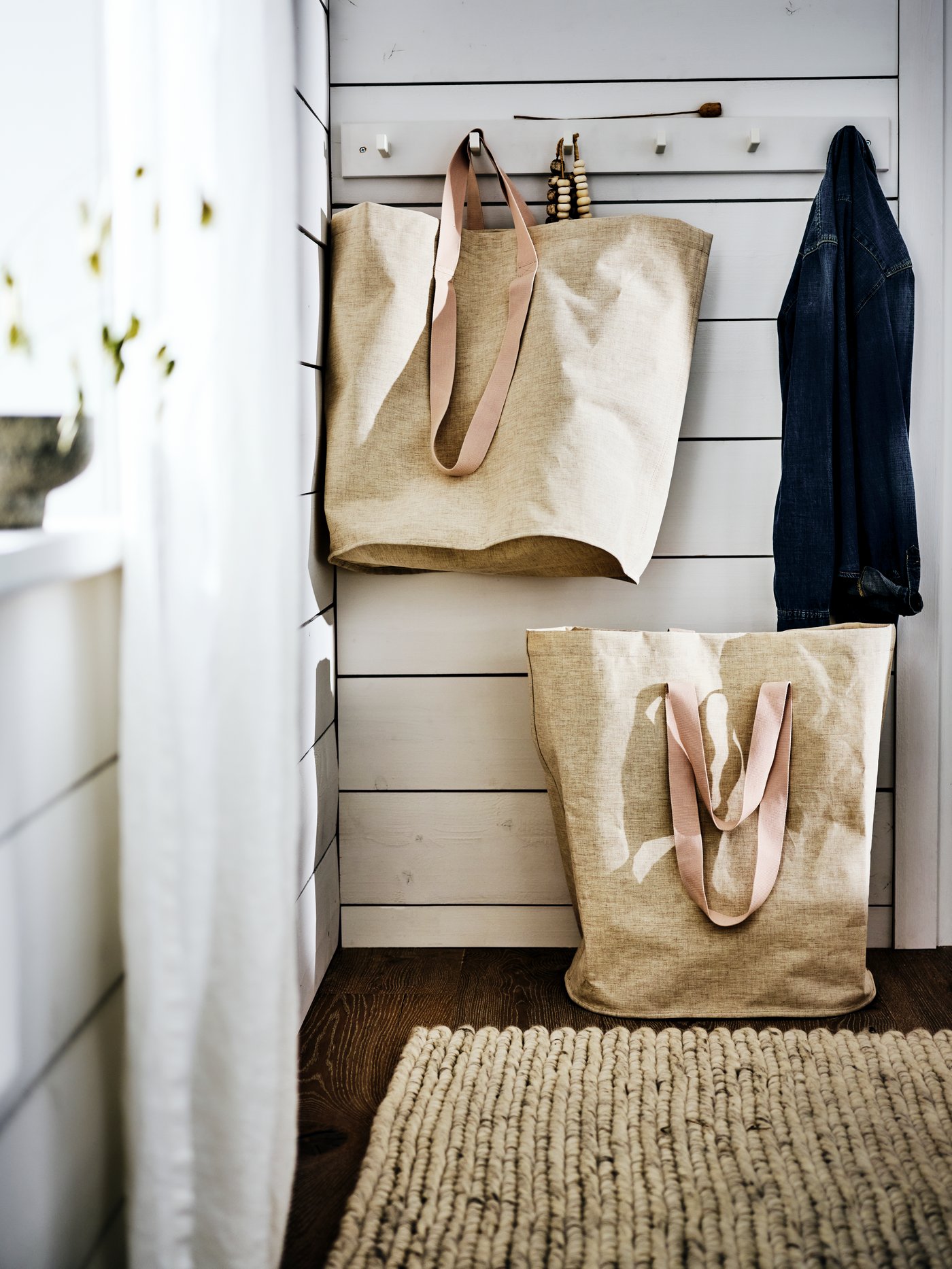 A white wood panelled wall with a row of hooks holding a large beige bag and a coat. A beige bag is below on the floor.