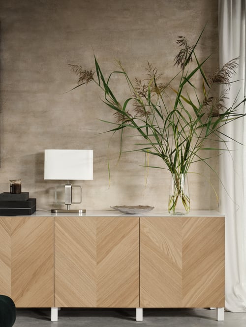 Three BESTÅ storage cabinets with herringbone-patterned fronts against a beige wall with a lamp and a glass vase on top.