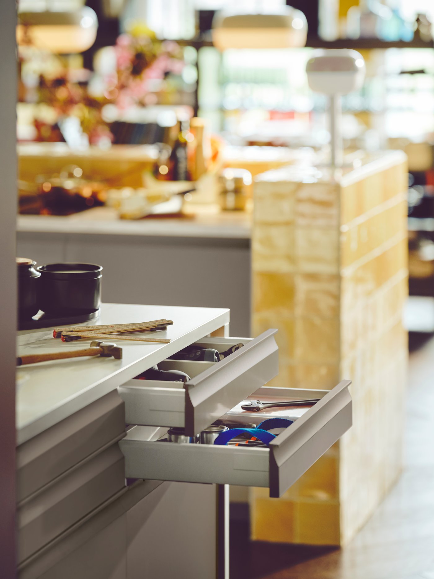 En öppen låda i en köksmiljö. Lådan innehåller olika redskap, inklusive saxar och andra köksverktyg. I bakgrunden finns en bänkskiva med föremål på den, och en trästruktur är också synlig.