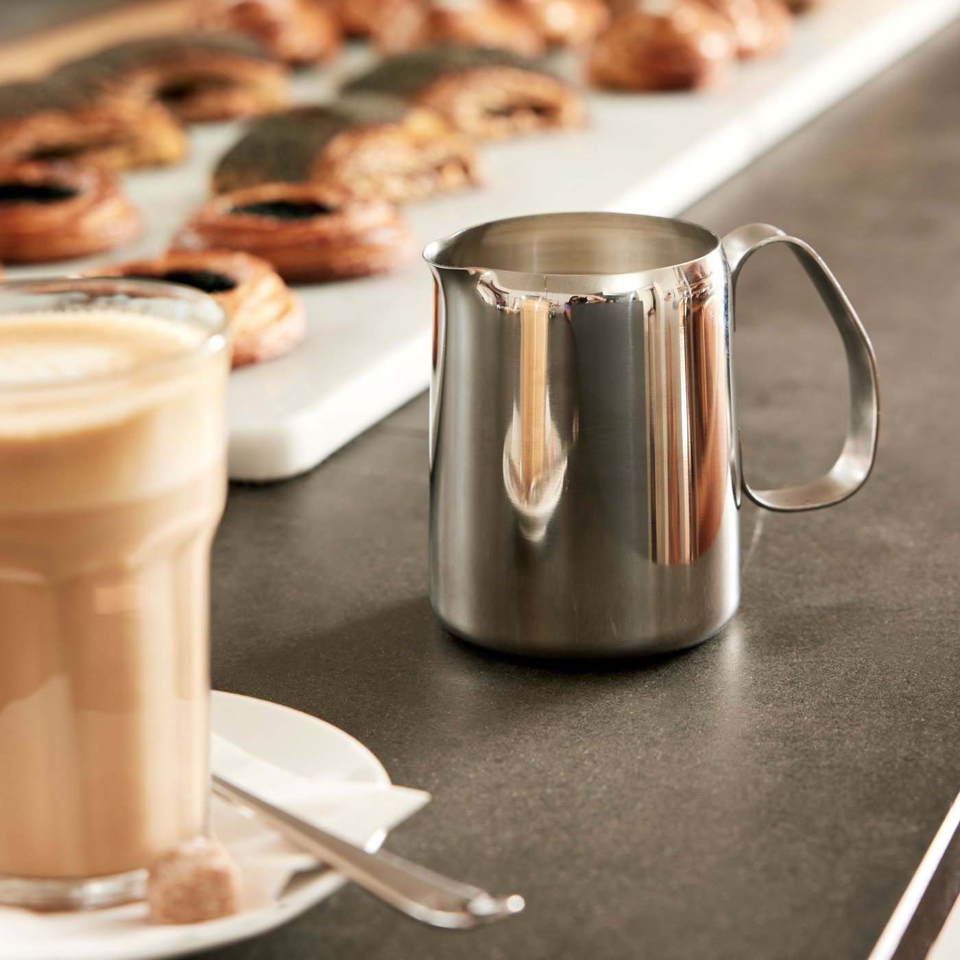 Gros plan d’un pichet à lait MATTLIG argent posé sur une table près d’un verre de café POKAL transparent.