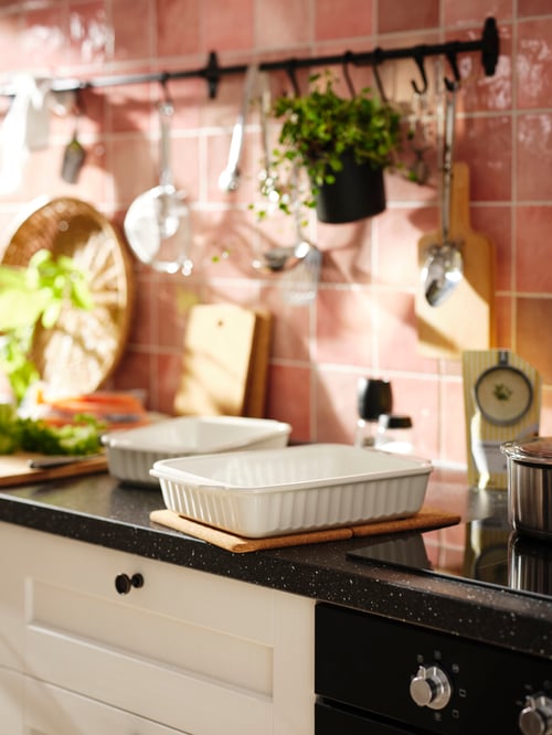 Close-up on kitchen counter with rectangular white stoneware oven dish on cork pot stand, rail with utensils in the back.