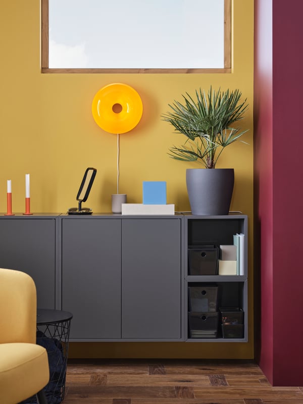 A colourful room with a grey EKET wall storage combination, a yellow wall lamp, a green plant and two orange candle holders.