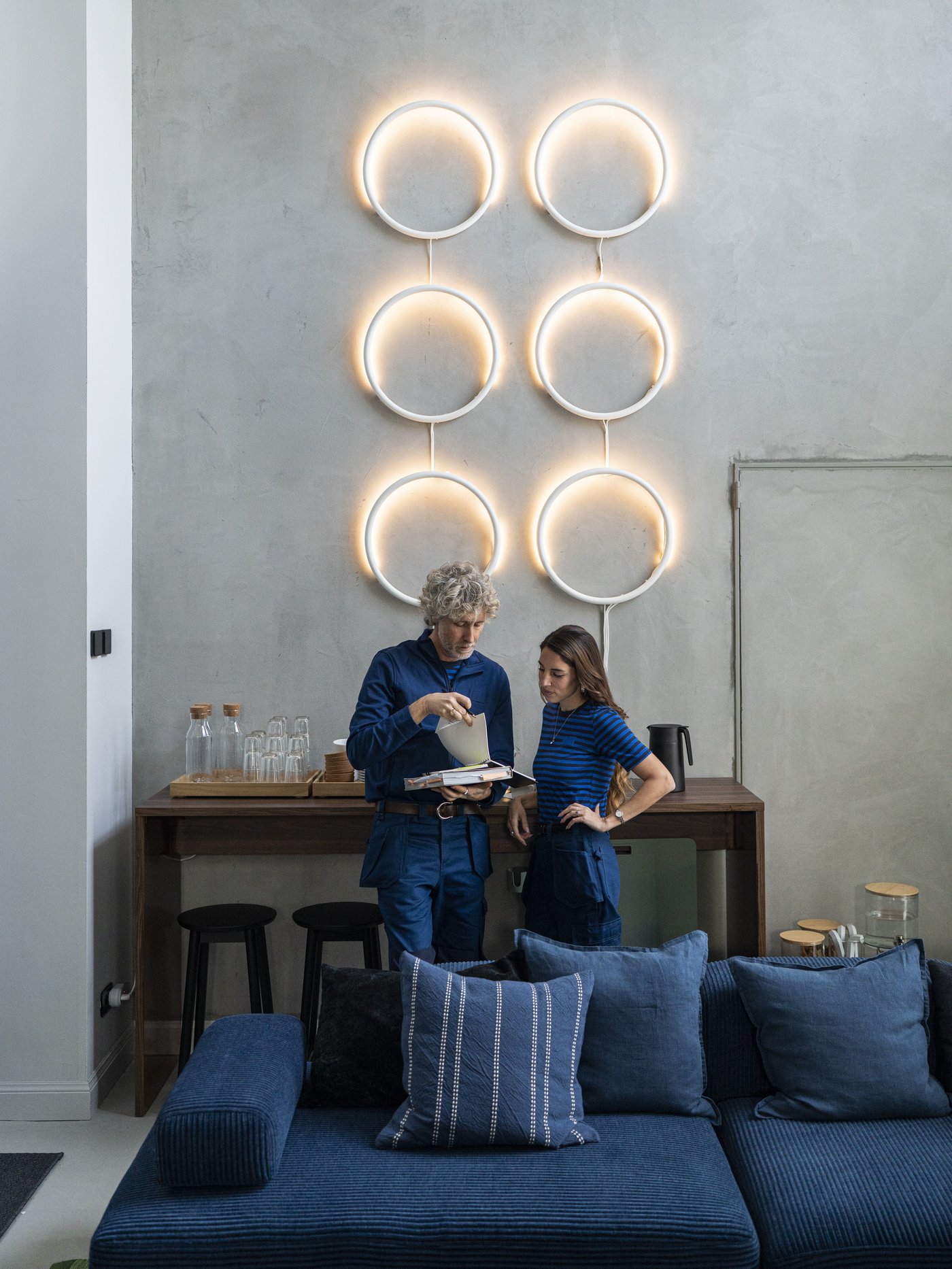 Soggiorno moderno con divano blu in primo piano, parete grigia decorata da sei lampade ad anello luminose e due consulenti IKEA che consultano dei documenti vicino a un tavolo consolle. 