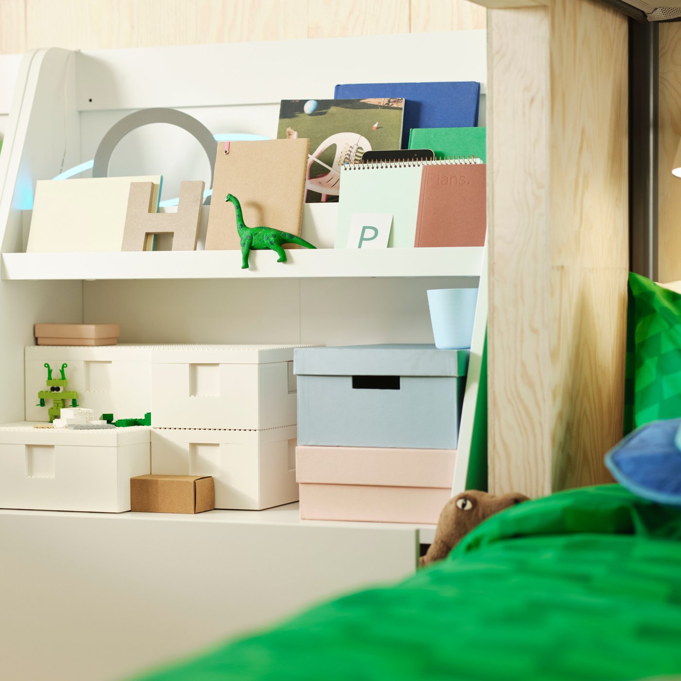 جزء من السرير به أقمشة باللون الأخضر المشرق وبجواره خزانة بيضاء وأرفف بيضاء عليها صناديق بألوان هادئة.