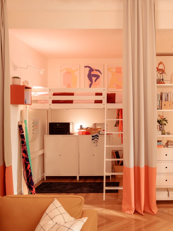 Pièce à l’éclairage chaleureux. À l’avant-plan sur la gauche, canapé beige avec coussin blanc. Au centre, lit superposé blanc surmontant des étagères. À droite, bibliothèque avec divers objets. Une lumière illuminant le plafond et des rideaux orange/blanc créent une atmosphère accueillante.