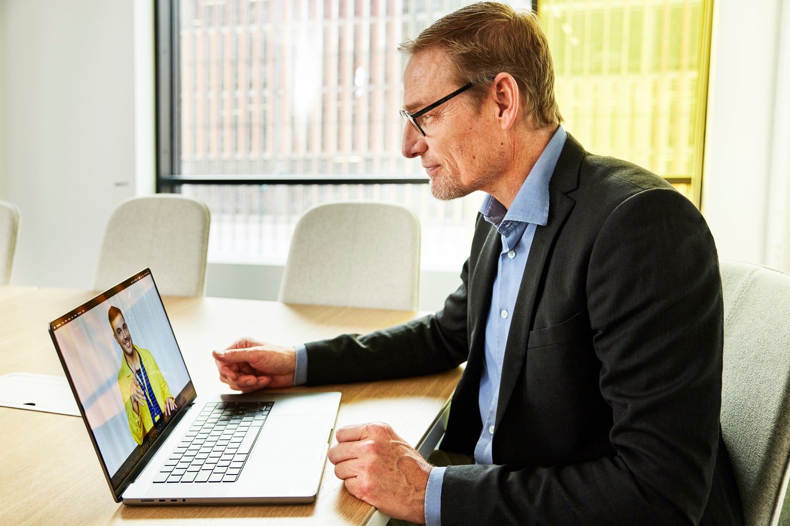 En mann i dress og briller sitter ved et møtebord og har et videomøte på laptopen sin.