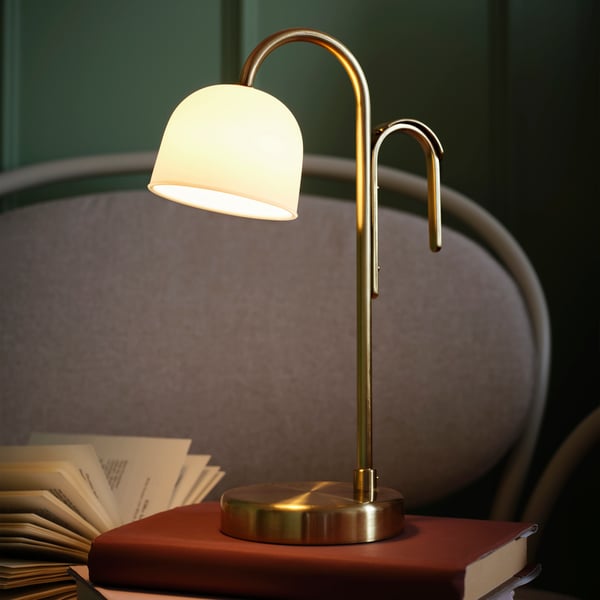 An opal white TVÅMASTAD LED portable lamp is elegantly placed on a stack of books on a cosy armchair.