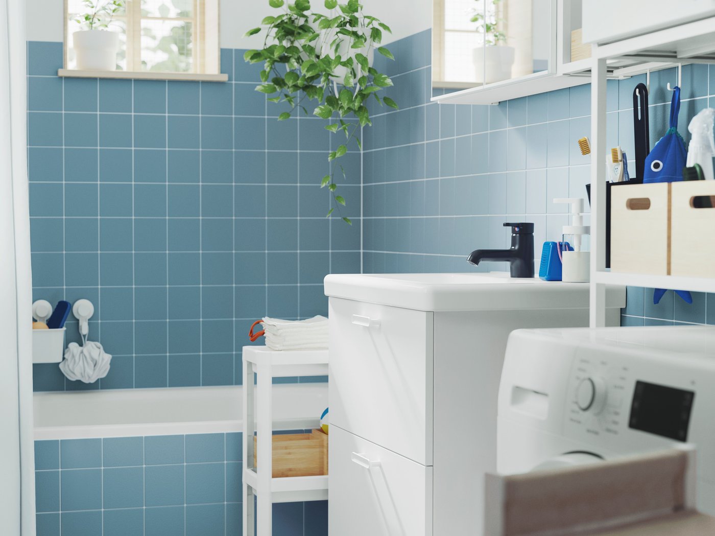 Modře vykachlíčkovaná koupelna s vanou, bílou spodní skříňkou ENHET, nástěnnou skříňkou se zrcadlovými dvířky a vozíkem VESKEN.