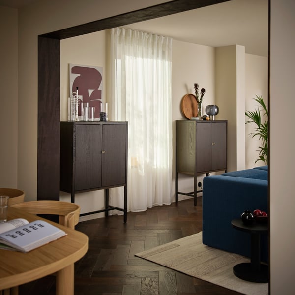 A living room with two dark-brown STOCKHOLM 2025 cabinets against a light wall and a window covered with sheer curtains.