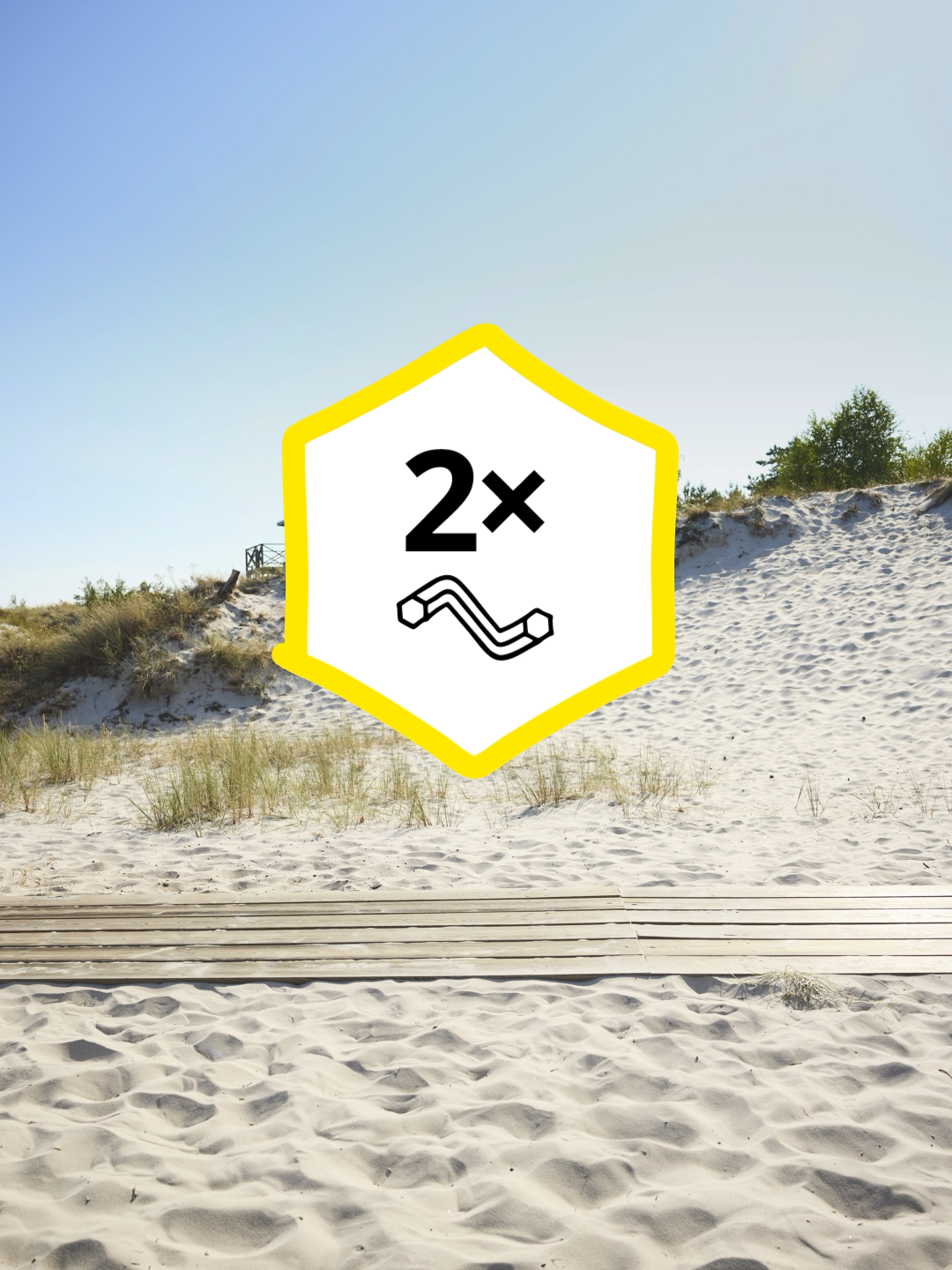 Dune di sabbia con passerella in legno sotto un cielo azzurro, con un simbolo esagonale giallo “2x” e un’icona a tubo