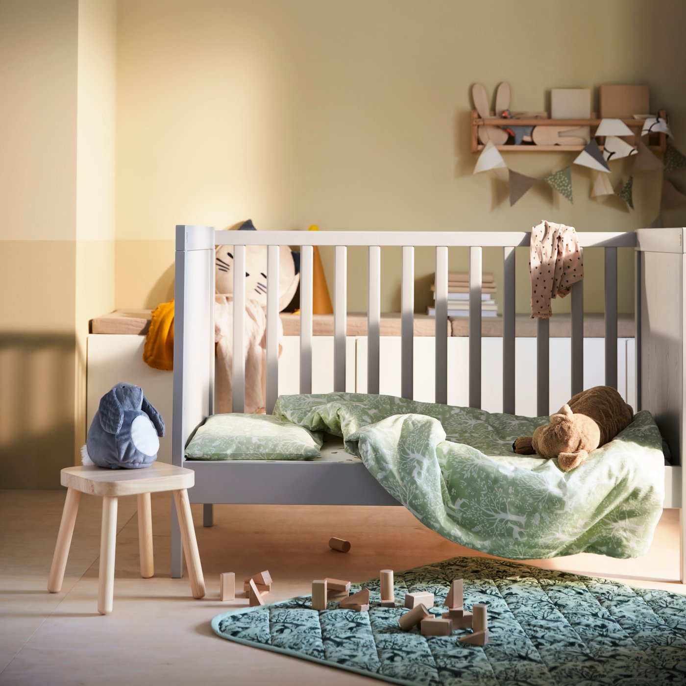 A TROLLDAM baby towel falling off a crib in a children's room. A stool sits next to it with a soft toy sitting on top of it. A rug and wooden toy blocks are on the floor sitting next to the crib as well.