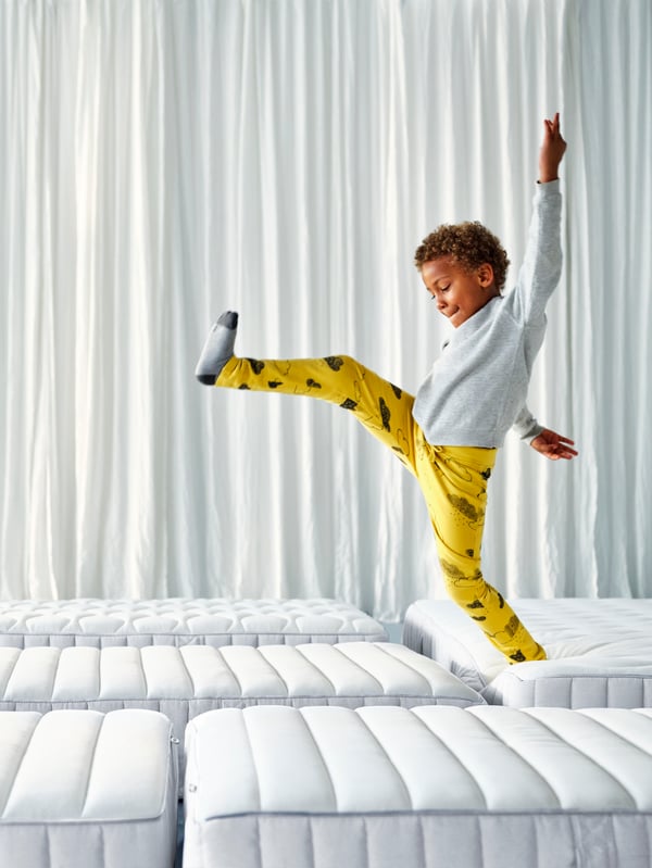 A young boy in bright yellow pyjamas trousers and a white top is jumping from one mattress to another in a white studio space.