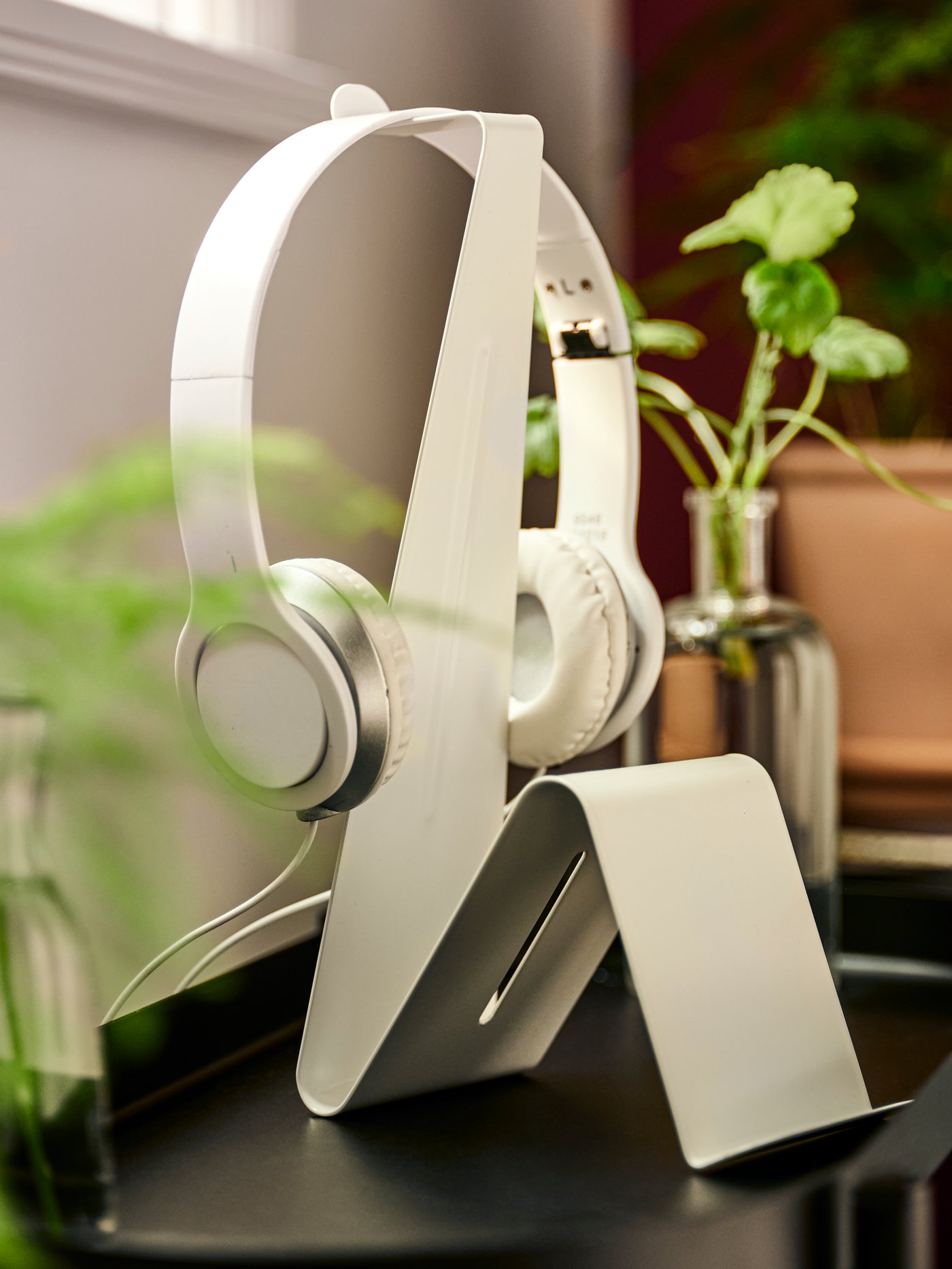 حامل سماعة رأس/كمبيوتر لوحي MÖJLIGHET أبيض مع زوج من سماعات الرأس البيضاء معلقة عليه، بين مزهريتين زجاجيين بها نباتات.