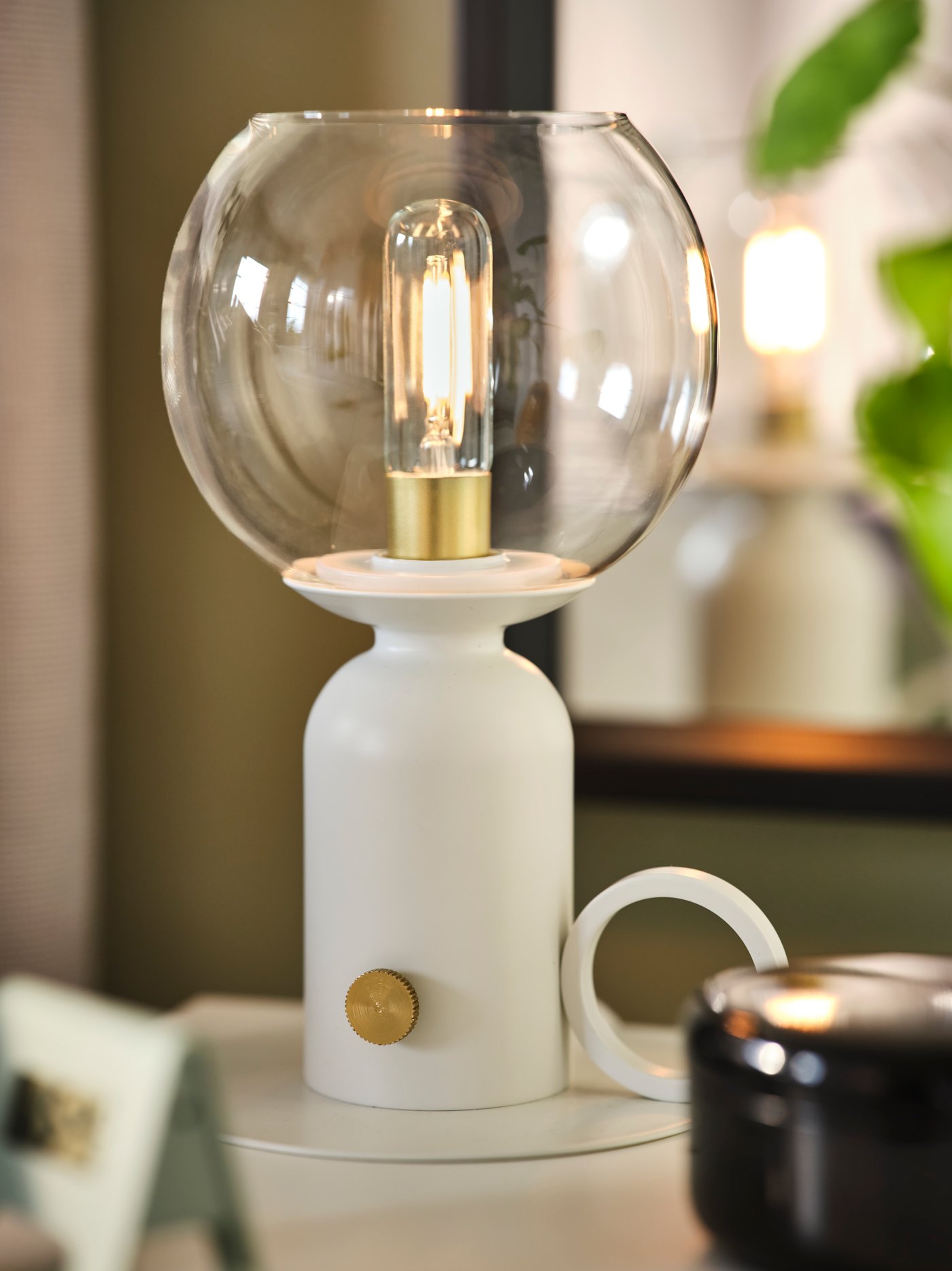 A lit, white ÅSKMULLER table lamp on a white surface with a green plant in the background.