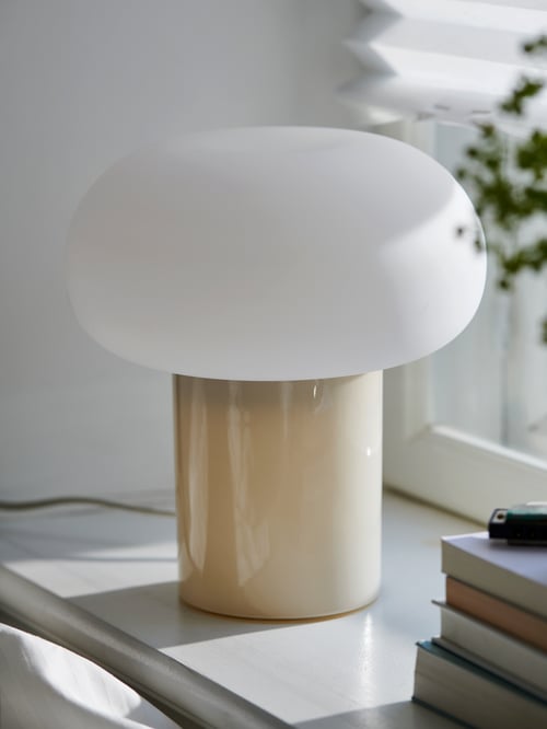 A DEJSA table lamp in a beige and opal-white glass combination stands next to a stack of books on a sunlit windowsill.